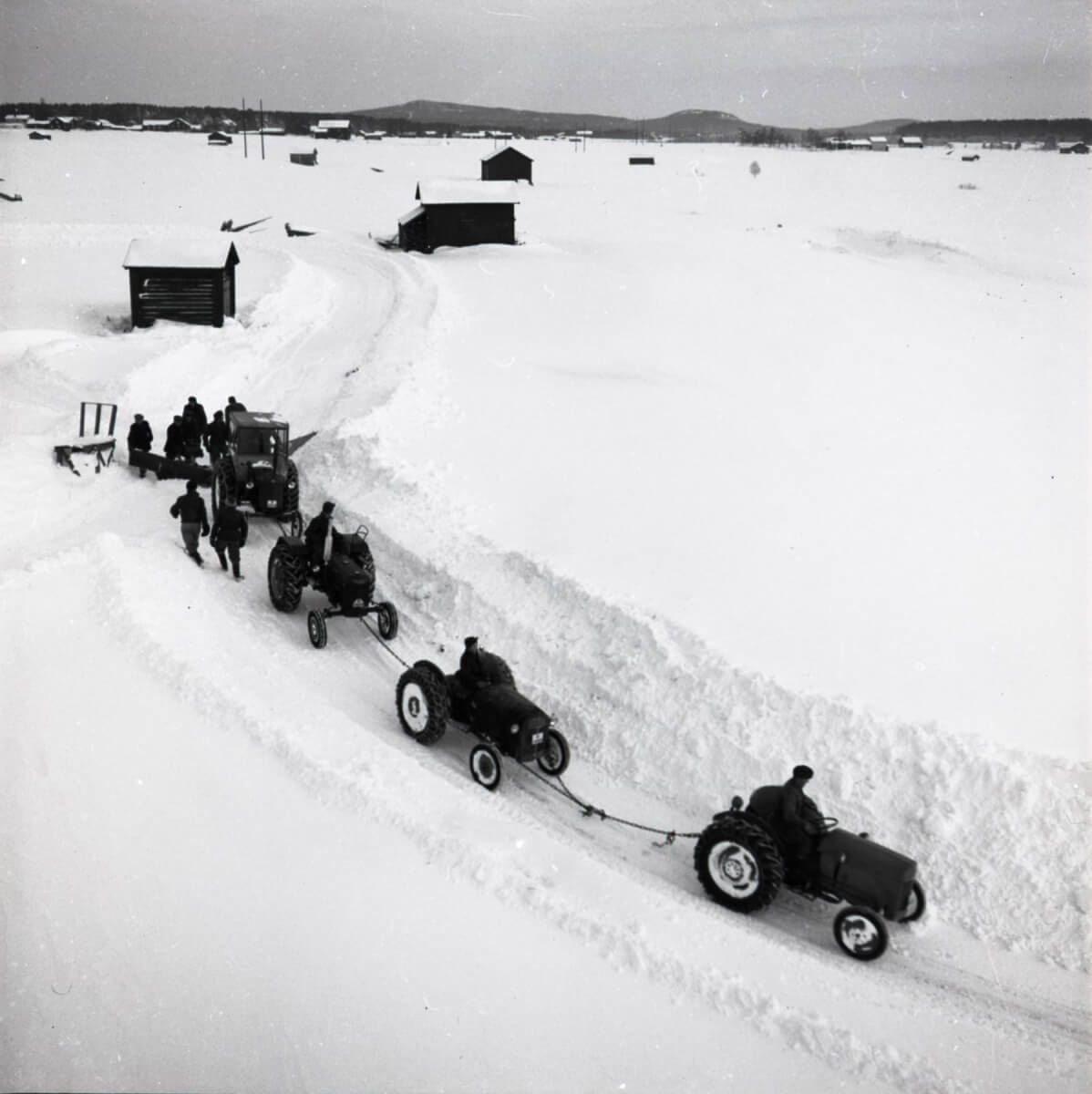 Fyra sammankopplade traktorer drar en plog i ett snöigt landskap.