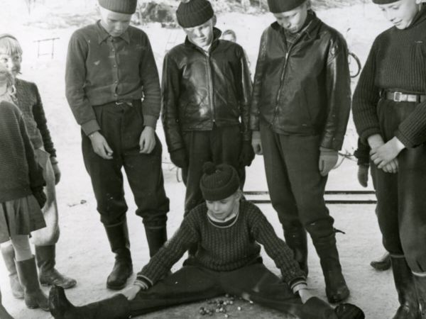 En grupp barn spelar kula. En pojke sitter ned och ramar in kulorna med sina ben.
