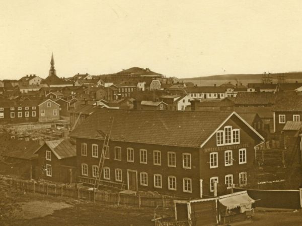 En mycket gammal bild av Hudiksvall med stora och små trähus som trängs längs gatorna. En stor träbyggnad i förgrunden har en skylt på gaveln där det står Molin & Co Snusfabrik. Man kan se Hudiksvallsfjärden och ett segelfartyg. Det gamla rådhuset sticker upp sin takspira till vänster. Bilden är tagen för 1879.