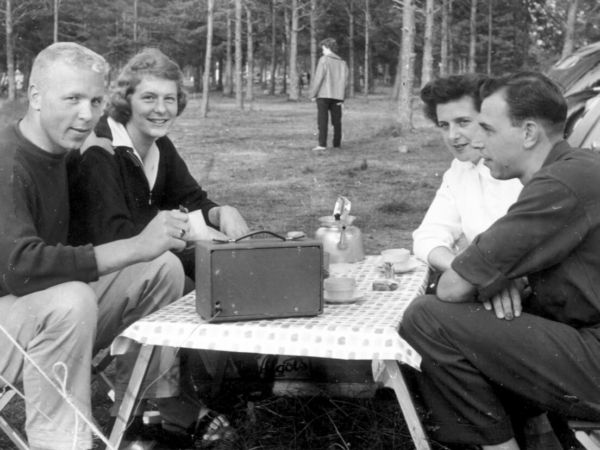 Fyra personer fikar runt ett fällbord och lyssnar på radio.