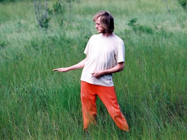 En man iförd orangea byxor och vit t-shirt står i gräset.